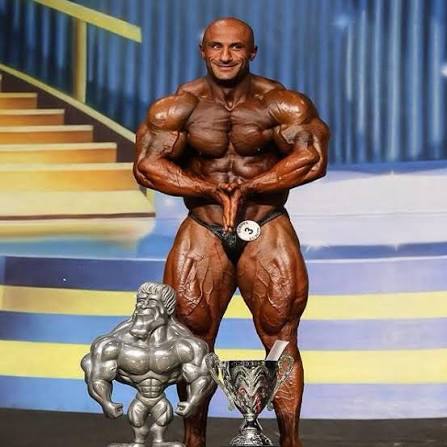 Moe Bannout posing onstage at an IFBB bodybuilding competition with the Europa Phoenix Pro trophies